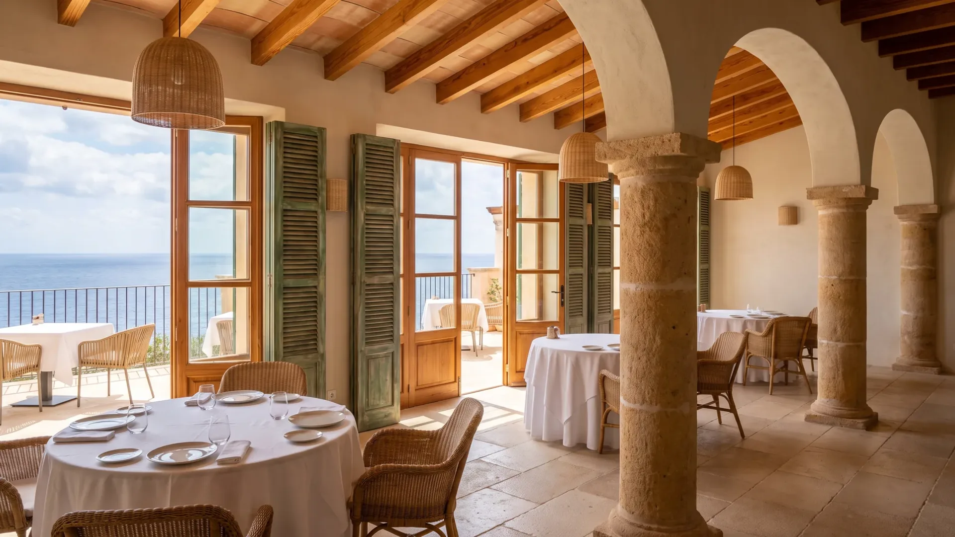 Hotel restaurant with tables laid in white linen, rattan chairs, stone columns and arches, and doors open to the sea