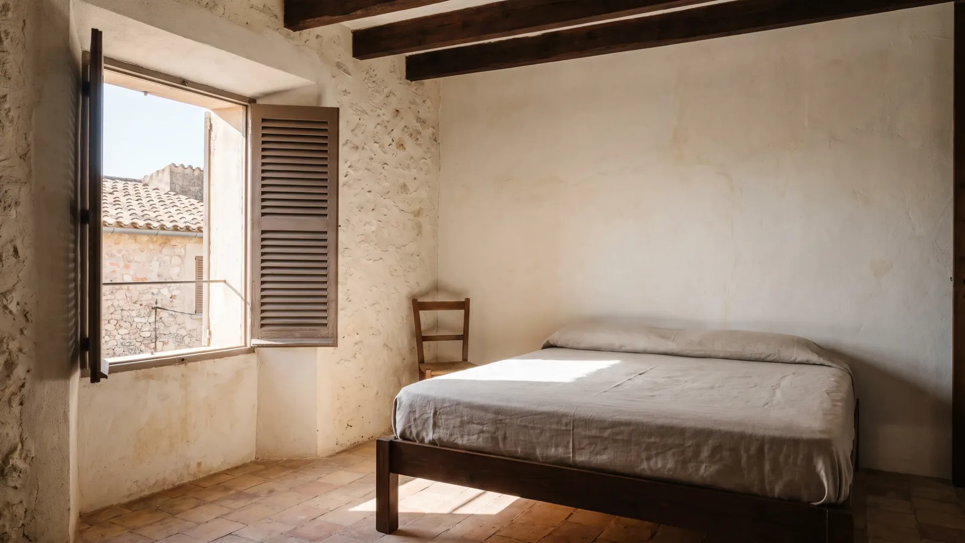 Bedroom in a stone house with a linen-covered bed, a timber chair and sunlight from a shuttered window on terracotta