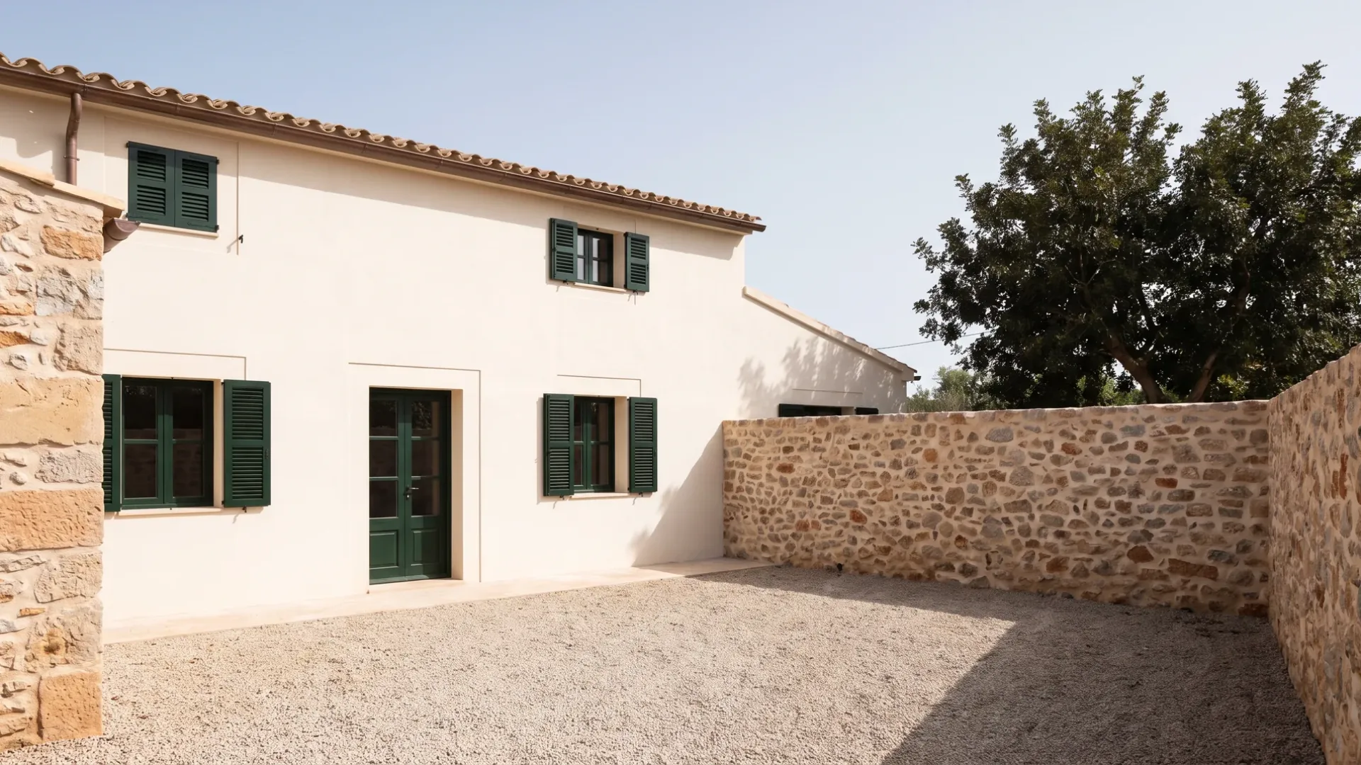 Village house in warm white render with dark green shutters, a gravel yard inside a repointed stone wall, a carob tree