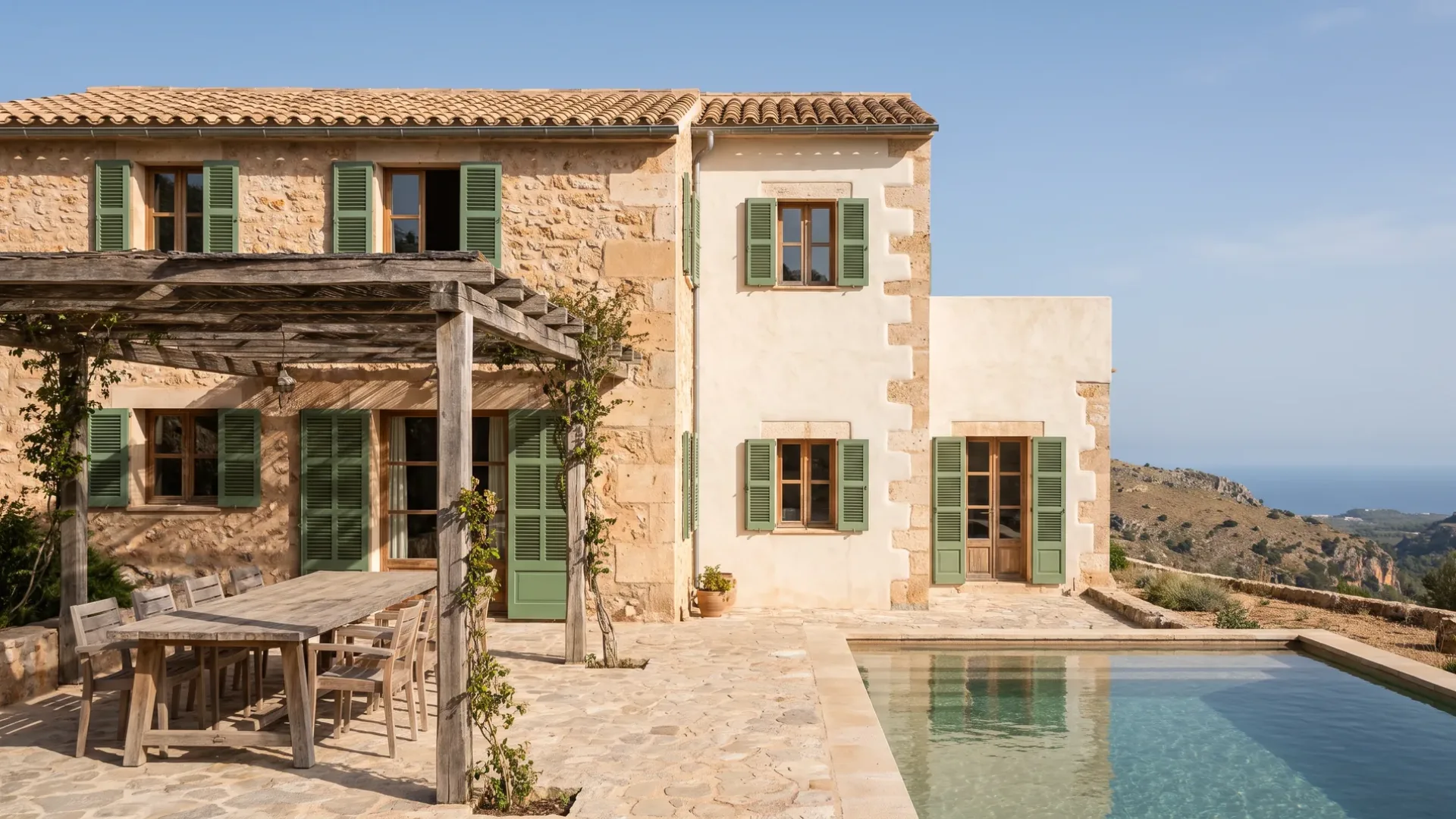 Stone villa on a hillside with green shutters, a pergola over a terrace table and a pool looking toward the sea