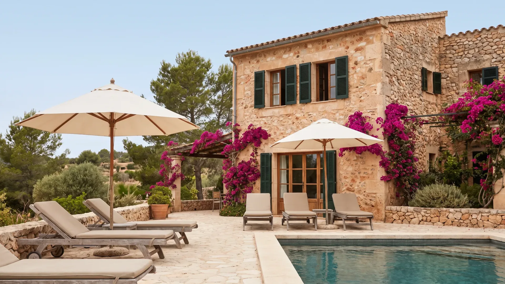 Litet stenhotell med gröna fönsterluckor och bougainvillea, solstolar och parasoll runt en pool på en stenterrass