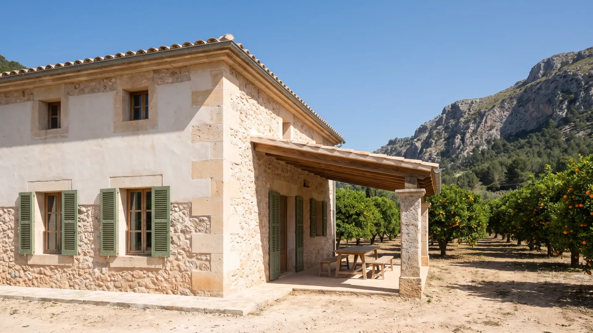 Kalkputsad finca av sten med gröna fönsterluckor och en veranda på en stenpelare, apelsinträd och en brant bergssida